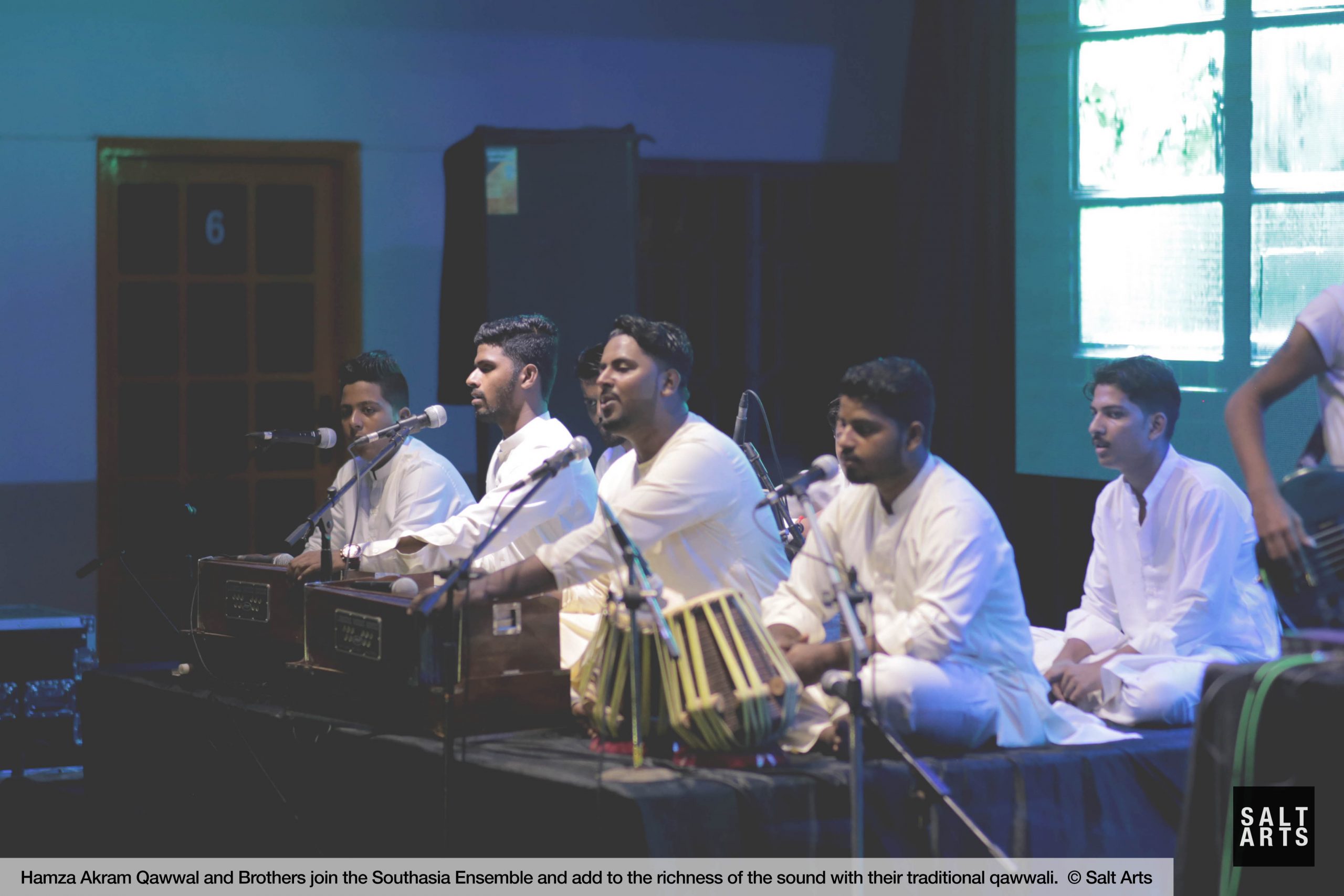 A group of male Indian classical musicians on a stage singing and playing their respective instruments