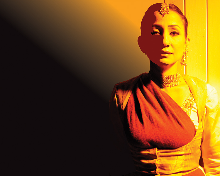 A woman lit in a bright yellow wash with gold earrings, a tikka and a red scarf across her chest