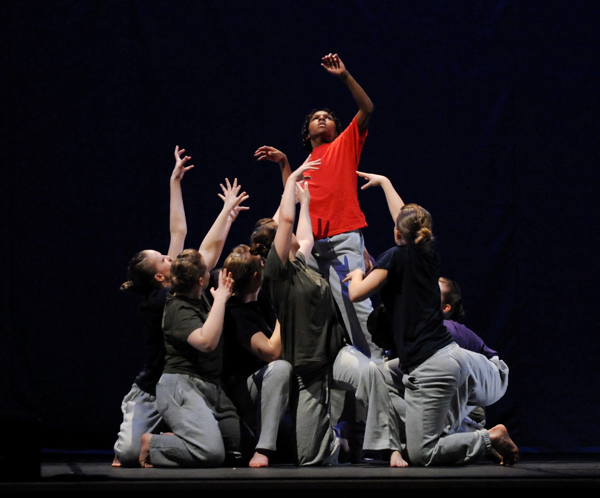 IDFb dance group performing with people in black and grey costumes at a lower level with a boy in a red top and grey joggers raised above them