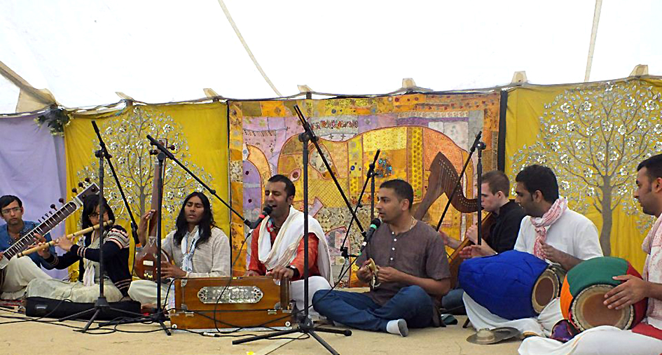 a group of musicians and vocalists sat singing with microphones in front of walls of tapestries