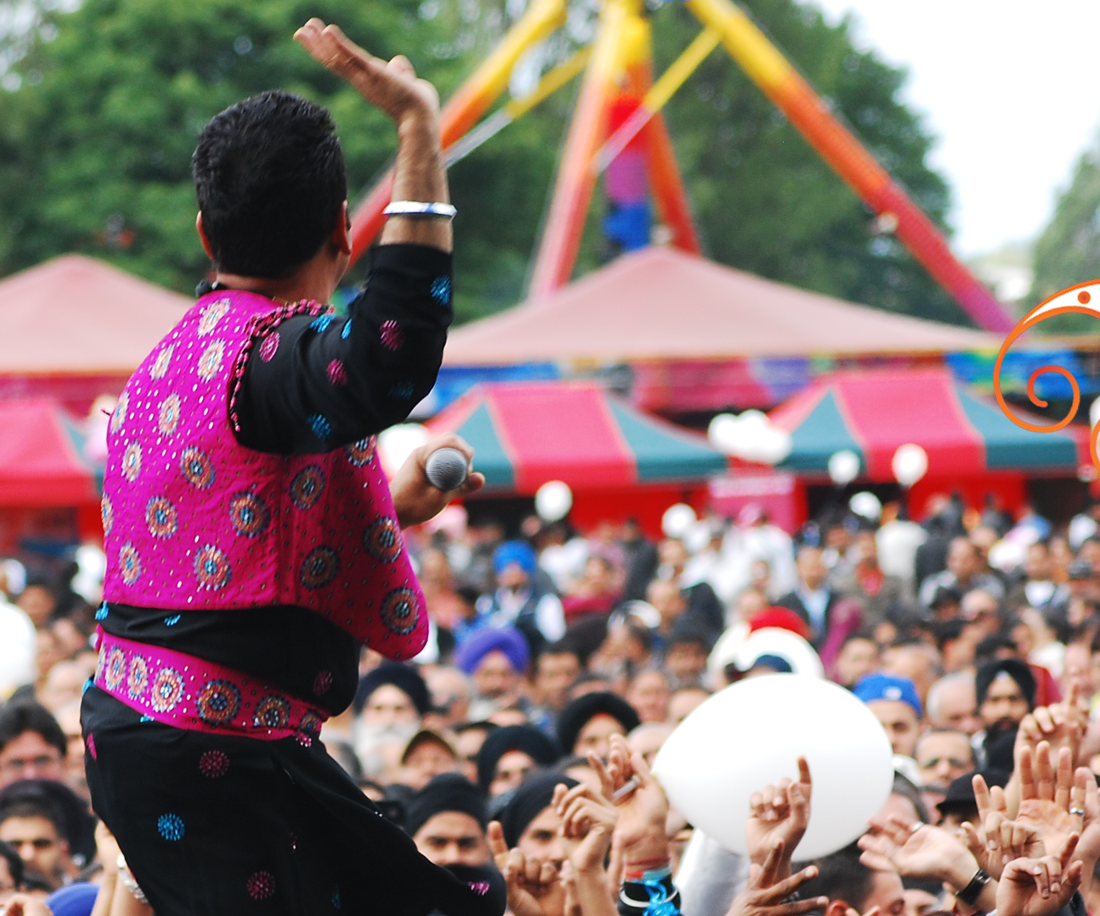 a man in a pink and black patterned outfit sits above a full crowd with his hand held up at a fair/ festival setting