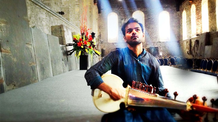 Soumik Datta in a stone building sat on a stage with a stringed instrument