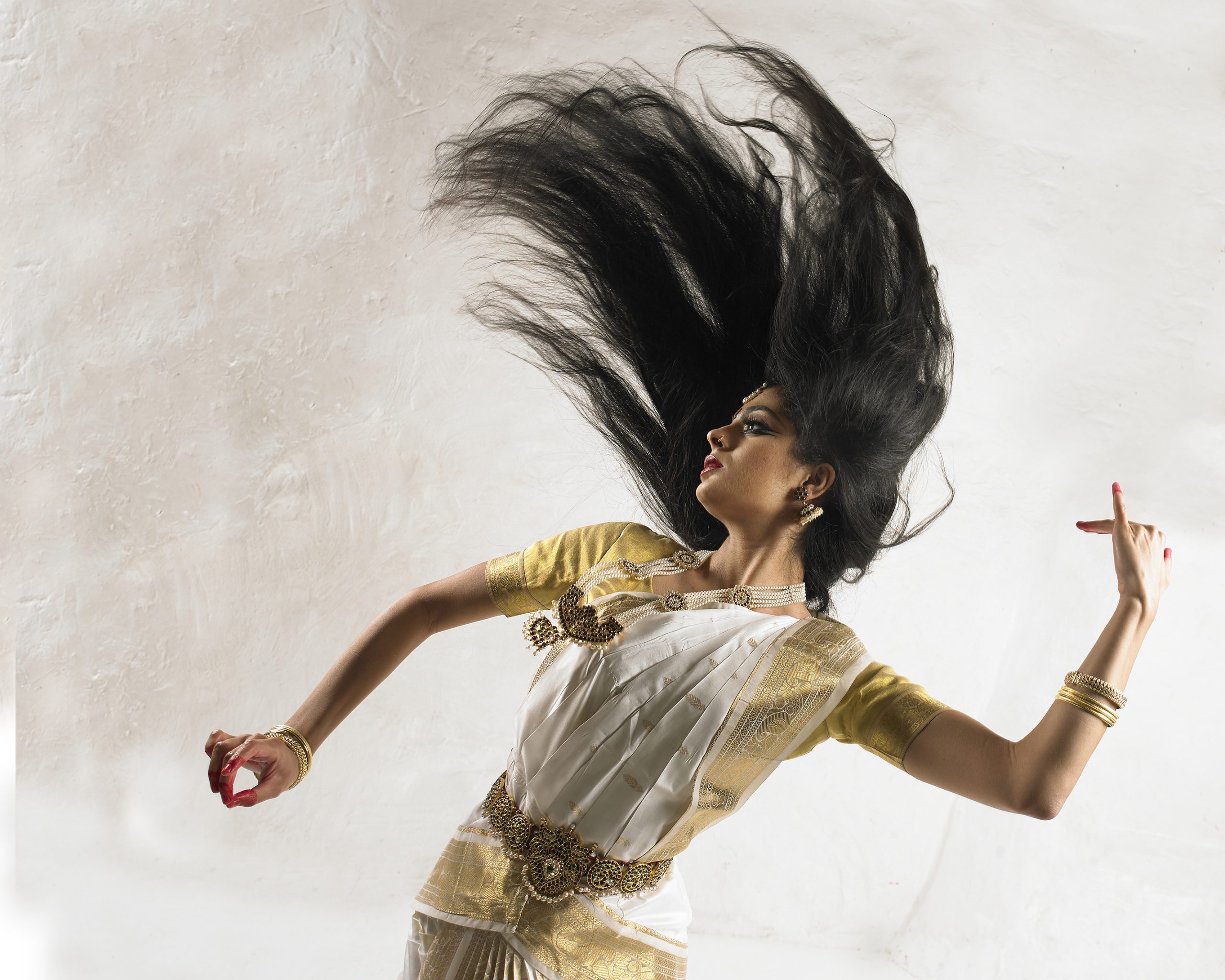 A close up on Anaya dancing in a white and gold Indian outfit with gold accessories mid-dance with her hair suspended in the air above her