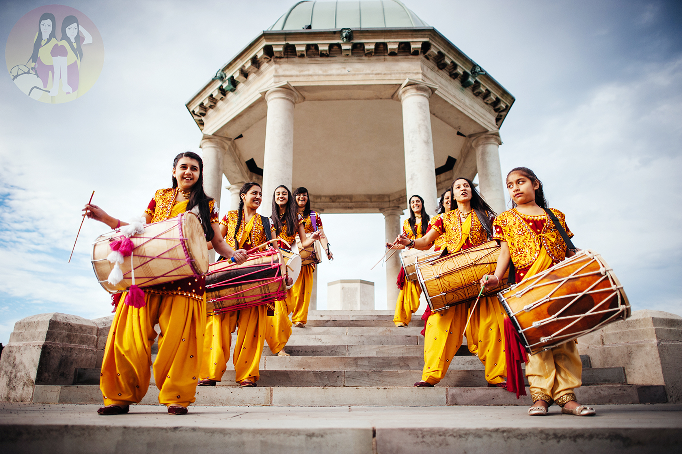 Under a gazebo monument, two rows of women and girls are dressed in orange with dhols held smiling and with different facial expressions