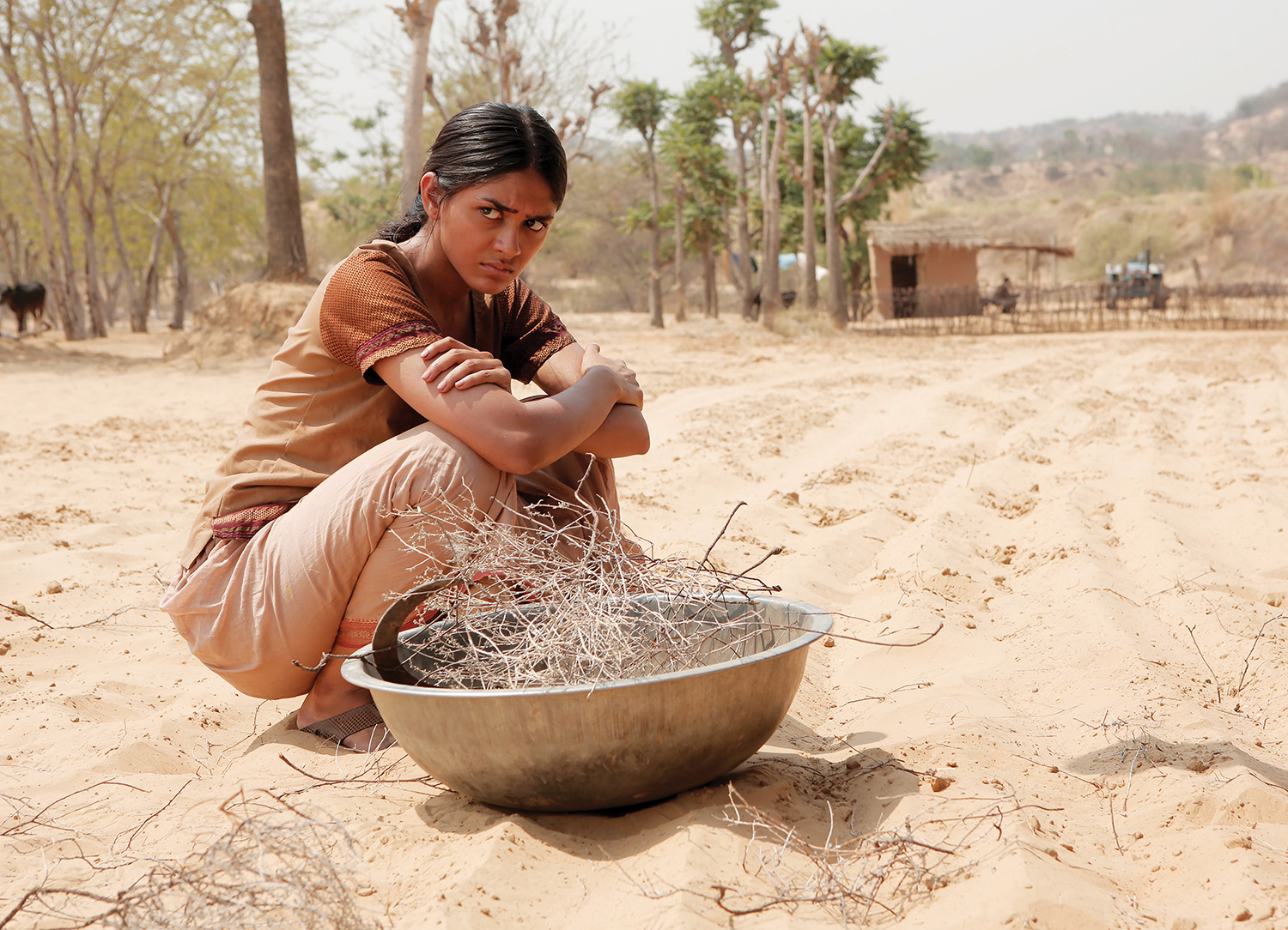 A girl crouched in the sand cross-armed with a metal bowl full of twigs, looking off upset