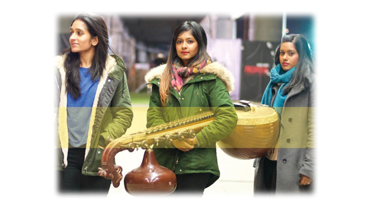Three women dressed in jackets and scarves in a line, the central figure holds a large stringed instrument