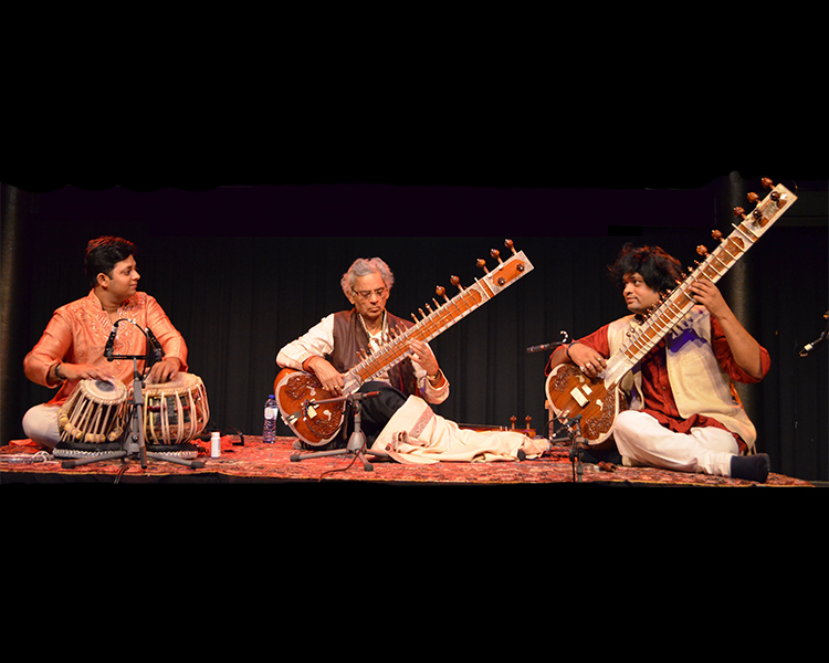 Shivnath Mishra music, three men one with tablas, two with stringed instruments on a stage with mics