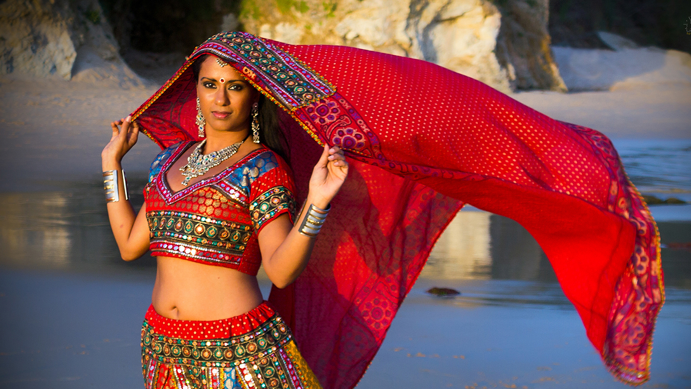 Sneha Mistra in a red lengha looking at the camera with her veil held over her head blowing in the wind against a water and rocky landscape