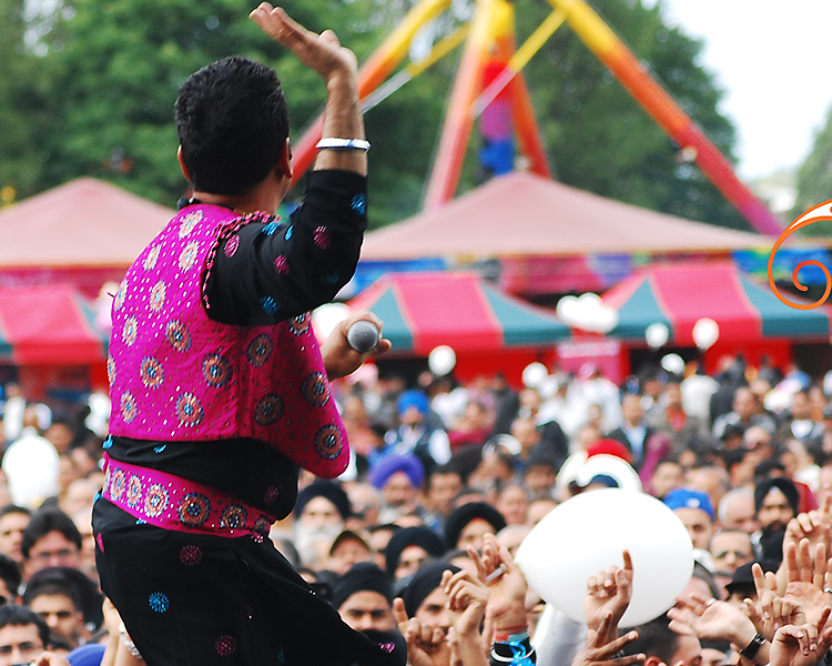 a man in a pink and black patterned outfit sits above a full crowd with his hand held up at a fair/ festival setting