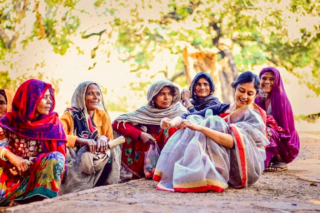 a group of women in saris sat on the ground laughing, and smiling